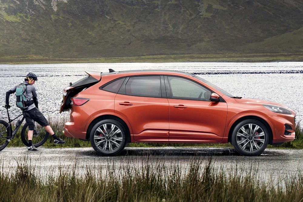 Une Ford Kuga orange vue de côté, garée près d'un lac, avec un cycliste chargeant du matériel dans le coffre ouvert.