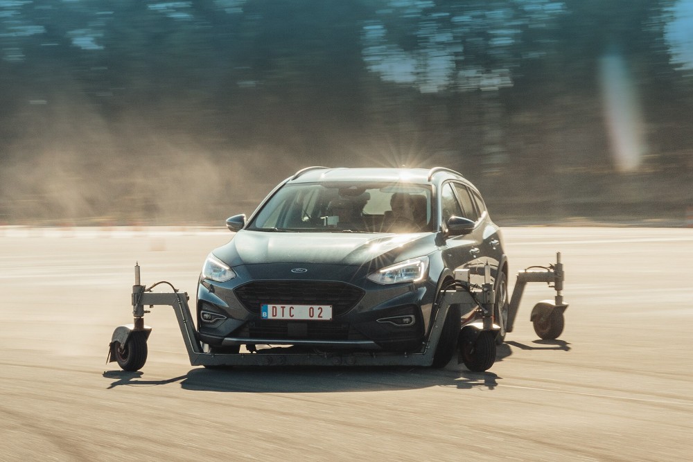 Une Ford Focus grise équipée d'un dispositif de test roulant sur une piste, avec de la poussière soulevée à l'arrière.