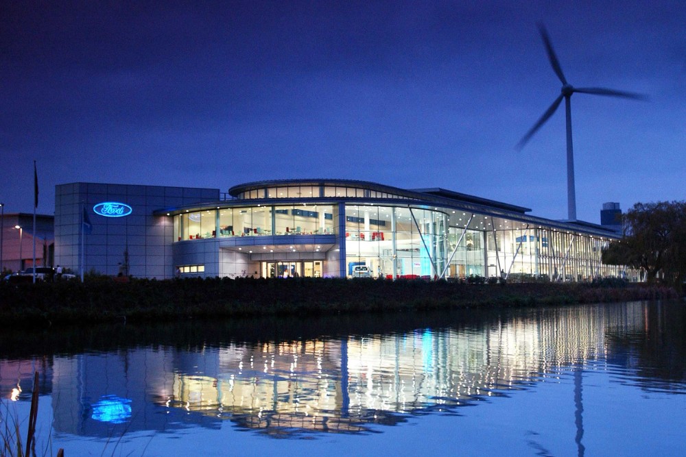 Un bâtiment Ford illuminé au crépuscule, avec une éolienne et son reflet dans l'eau au premier plan.