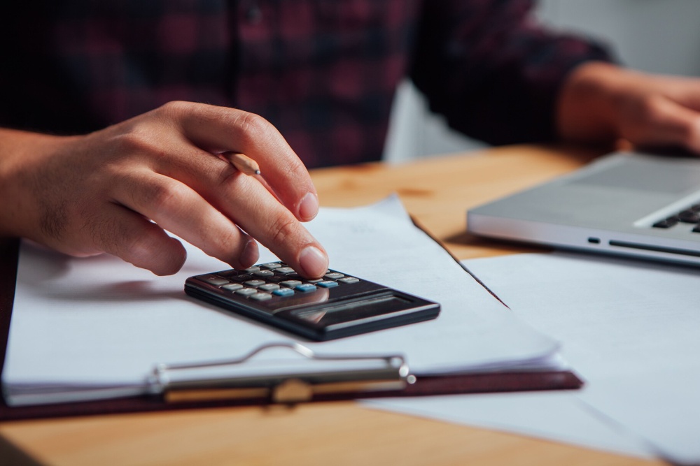 Une main tape sur une calculatrice posée sur des documents, un ordinateur flou à l'arrière-plan.