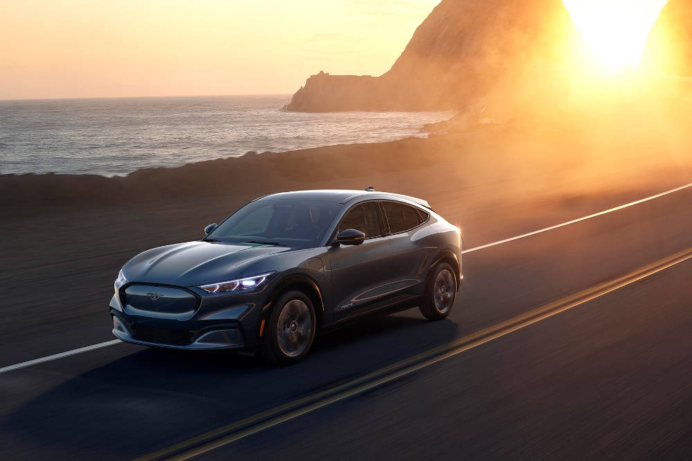 Une voiture électrique Ford Mustang Mach-E roule sur une route côtière au coucher du soleil.