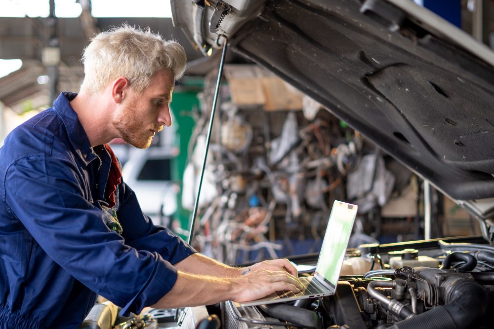 Un technicien en uniforme bleu utilise un ordinateur portable tout en travaillant sur un moteur de voiture capot ouvert.