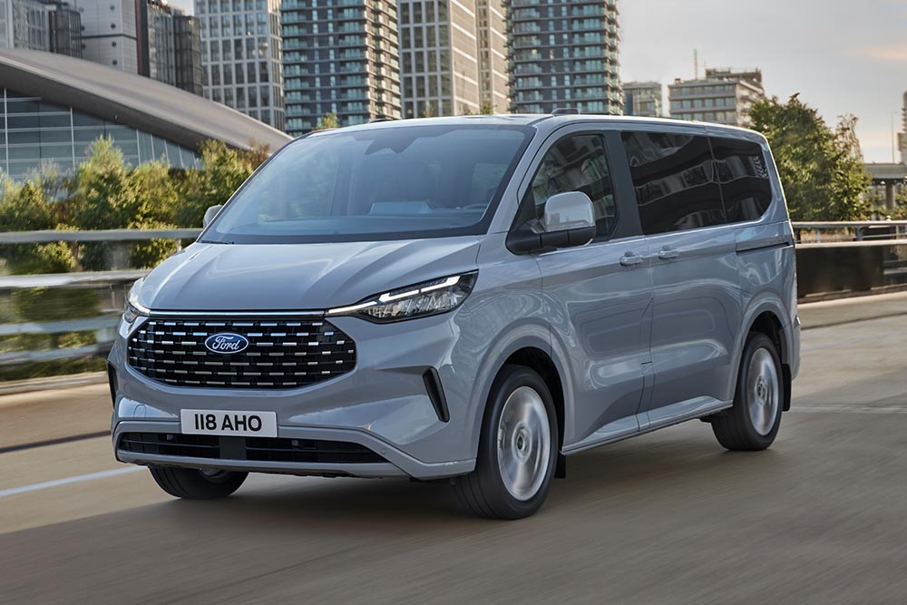 Un Ford Tourneo Custom argenté roule sur une route en ville.