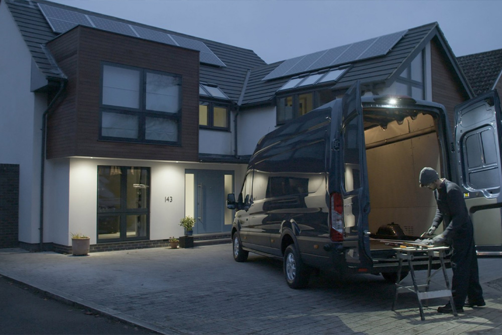 Un menuisier travaille derrière un fourgon Ford Transit aux portes arrière ouvertes, éclairé par le plafonnier LED.