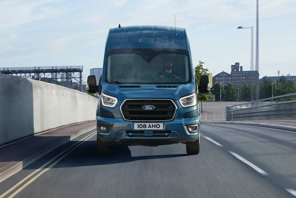Vue frontale d’un fourgon Ford Transit bleu circulant sur une route. 