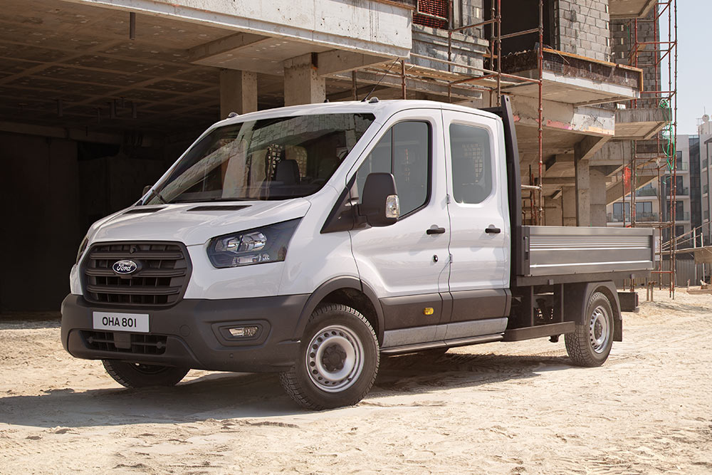 Un Ford Transit Custom blanc à châssis-cabine avec plateau est garé sur un chantier.
