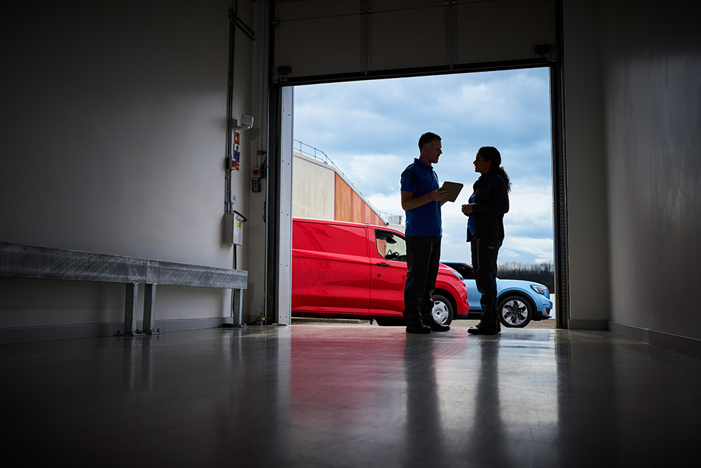 Deux personnes parlant devant une porte de garage, avec un fourgon Ford rouge et un SUV Ford bleu stationnés à l’extérieur.